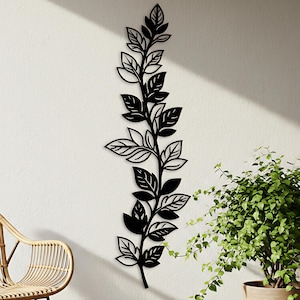 Pode incluir: Arte de parede em metal preto que retrata um ramo de folhas estilizado. A obra de arte é montada em uma parede branca, com uma cadeira de vime e uma planta em vaso ao fundo. As folhas têm detalhes recortados intrincados.