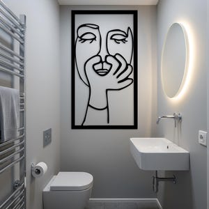 May include: A modern bathroom with a minimalist design. Features a white toilet, sink, and a large black framed wall art with a line drawing of a face. A heated towel rack and a round mirror with a glowing edge complete the space.
