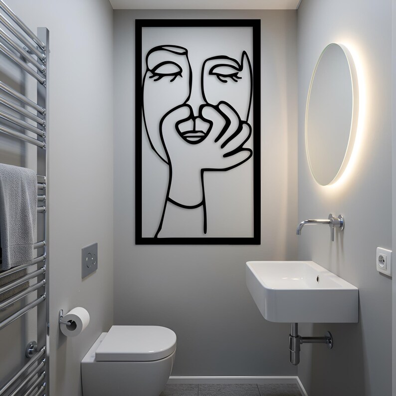 May include: A modern bathroom with a minimalist design. Features a white toilet, sink, and a large black framed wall art with a line drawing of a face. A heated towel rack and a round mirror with a glowing edge complete the space.