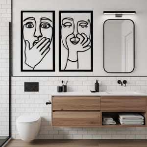 May include: Two framed wall art pieces with black outlines of faces, one with a hand over the mouth and the other with a hand near the mouth. The art hangs above a wooden vanity with a black-framed mirror and a white sink.