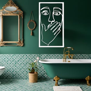 May include: A bathroom with a green and white patterned tile floor and a dark green wall. A white framed line art of a face with a hand over the mouth hangs on the wall. A gold framed mirror and a gold faucet are also visible.