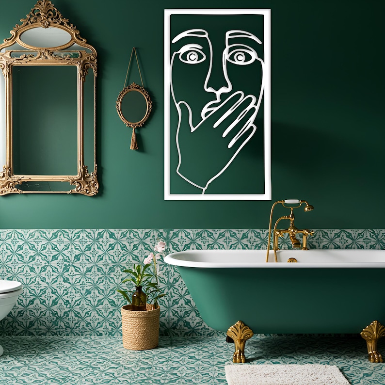 May include: A bathroom with a green and white patterned tile floor and a dark green wall. A white framed line art of a face with a hand over the mouth hangs on the wall. A gold framed mirror and a gold faucet are also visible.