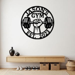 May include: Black metal wall art with the text "JASON'S GYM" and "EST. 2023" in a circular design. The design features a hand holding a barbell. The wall art is mounted on a white wall above a wooden console.