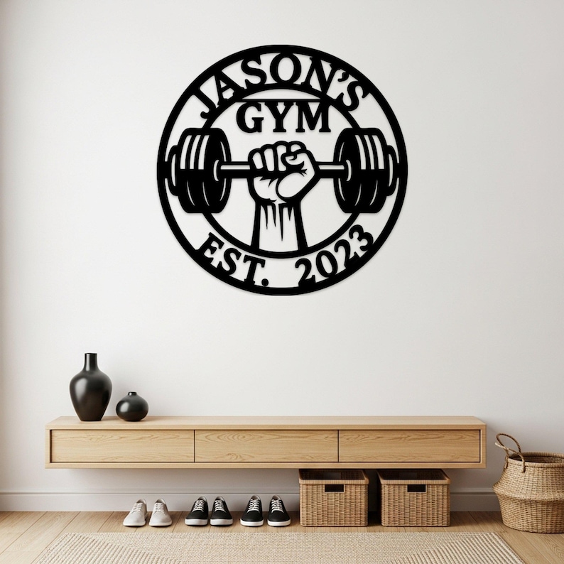 May include: Black metal wall art with the text "JASON'S GYM" and "EST. 2023" in a circular design. The design features a hand holding a barbell. The wall art is mounted on a white wall above a wooden console.