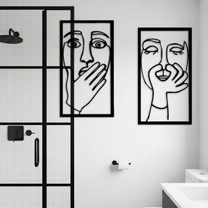 May include: Two framed wall art pieces with black line drawings of faces. One face has a hand covering the mouth, the other has a hand near the mouth. The art is displayed in a modern bathroom setting with a black shower door.