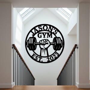 May include: Black metal wall art with the text "JASON'S GYM EST. 2023" in a circular design. The design features a hand holding a barbell. The art is mounted on a white wall above a staircase.