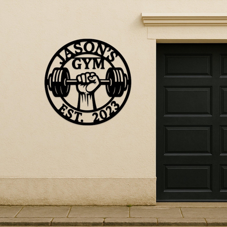 May include: Black metal wall art with the text "JASON'S GYM EST. 2023" in a circular design. A hand is lifting a barbell in the center of the circle. The art is mounted on a beige wall next to a black door.