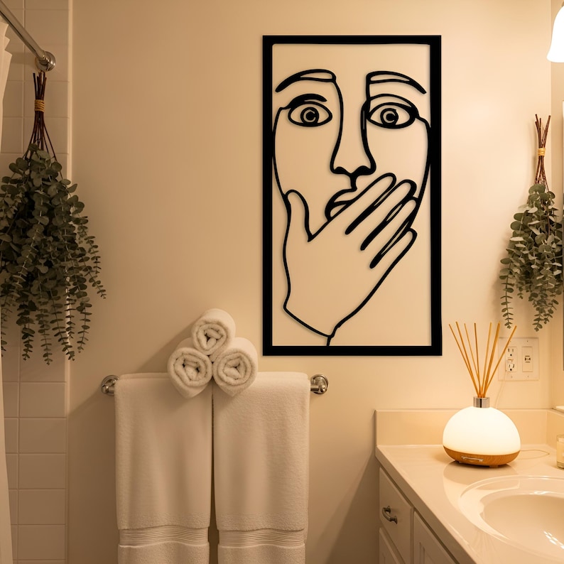 May include: A black framed wall art piece with a minimalist line drawing of a face, hand covering the mouth. Below, white rolled towels hang on a chrome towel rack. A small, round, white and wood-toned diffuser sits on the counter.