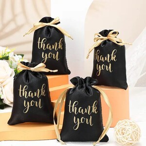 May include: Four black satin gift bags with gold "thank you" script. Each bag is tied with a gold ribbon. The bags are arranged on a light-colored surface, suggesting a celebratory or gifting theme.