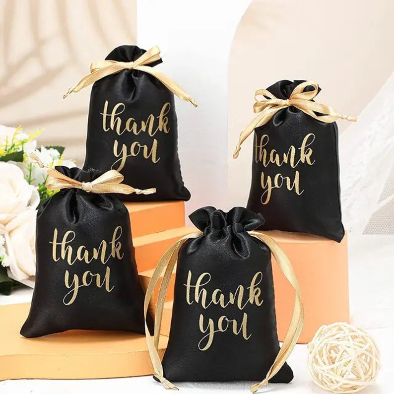 May include: Four black satin gift bags with gold "thank you" script. Each bag is tied with a gold ribbon. The bags are arranged on a light-colored surface, suggesting a celebratory or gifting theme.