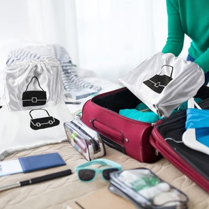 May include: White satin travel bags with a black handbag graphic are displayed near a red suitcase. Other travel accessories include sunglasses, a clear cosmetic bag, and a pen. The bags are being packed for a trip.