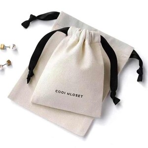 Puede incluir: Una bolsa de algodón blanco con cordones de ajuste negros y el texto "COOOI MLOSET" impreso en la parte delantera.