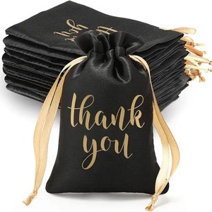 May include: Black satin drawstring gift bags with gold lettering that reads "thank you." The bags have gold satin ribbons for closure. A stack of similar bags is visible in the background.