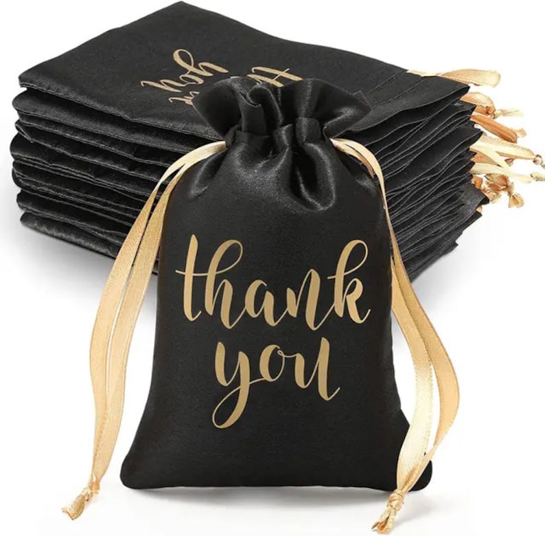 May include: Black satin drawstring gift bags with gold lettering that reads "thank you." The bags have gold satin ribbons for closure. A stack of similar bags is visible in the background.