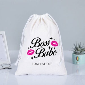 Puede incluir: Una bolsa de algodón blanco con cordón de ajuste, con el texto "Boss Babe" en letras negras y labios rosas con purpurina. El texto "Hangover Kit" está impreso debajo del texto principal.