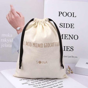 Puede incluir: Bolsa con cordón de color crema con cordones negros. La bolsa presenta el texto "IL MIO PRIMO GIOCATTOLO" en marrón, junto con la marca "SOINA". El fondo incluye texto y una vista parcial de una persona.