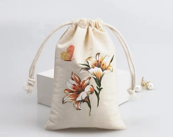 May include: A white cotton drawstring bag with a floral print featuring orange and white lilies and a small butterfly. The bag has a white background and the flowers are in shades of orange, white, and green.