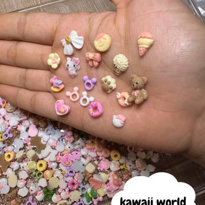 Pu&ograve; includere: Una variet&agrave; di ciondoli in miniatura colorati, tra cui una ciambella, un orsacchiotto, un personaggio Hello Kitty e un fiocco. I ciondoli sono sparsi su una mano e una superficie ricoperta di ciondoli pi&ugrave; piccoli.