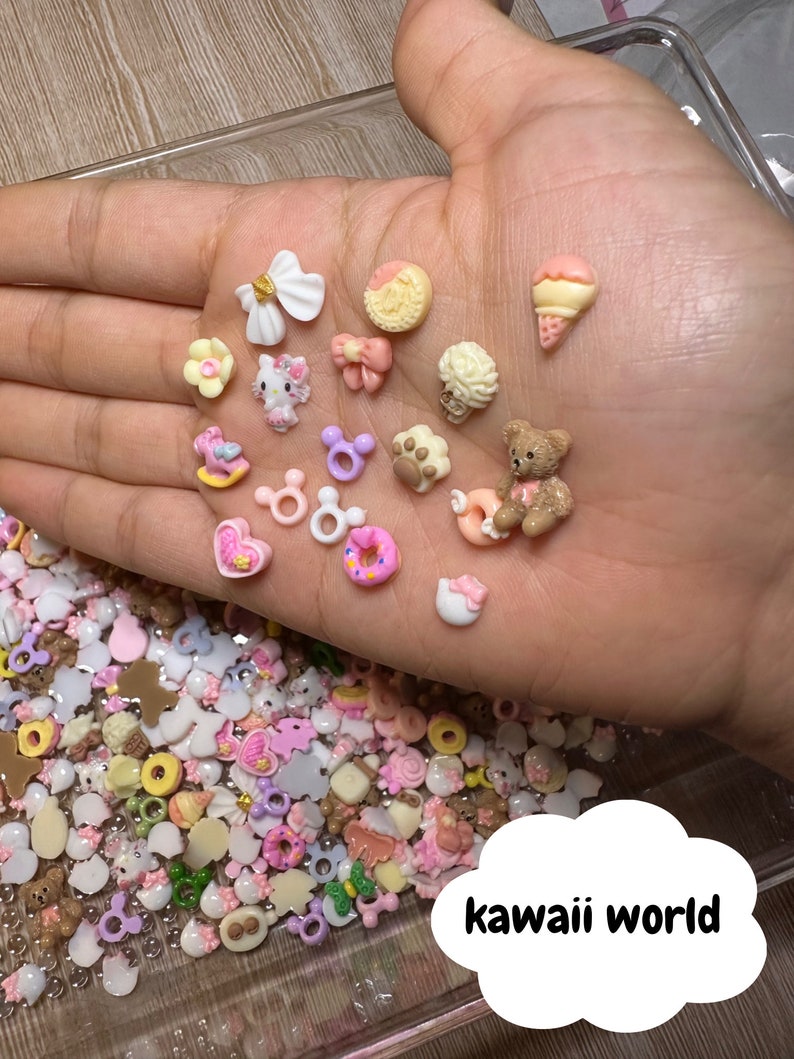 Pu&ograve; includere: Una variet&agrave; di ciondoli in miniatura colorati, tra cui una ciambella, un orsacchiotto, un personaggio Hello Kitty e un fiocco. I ciondoli sono sparsi su una mano e una superficie ricoperta di ciondoli pi&ugrave; piccoli.
