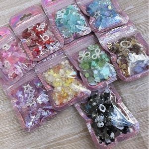 May include: A collection of small, iridescent pink zipper bags filled with various decorative embellishments. The bags contain an assortment of colorful beads, flowers, and charms in shades of red, blue, green, yellow, and purple, perfect for crafting and jewelry making.