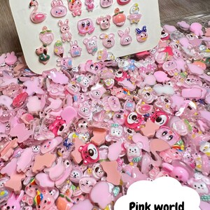 Pu&ograve; includere: Un vasto assortimento di ciondoli in plastica rosa e trasparente con vari animali e personaggi dei cartoni animati. I ciondoli sono sparsi su una superficie con un cartoncino bianco sullo sfondo che mostra una selezione pi&ugrave; piccola degli stessi ciondoli.