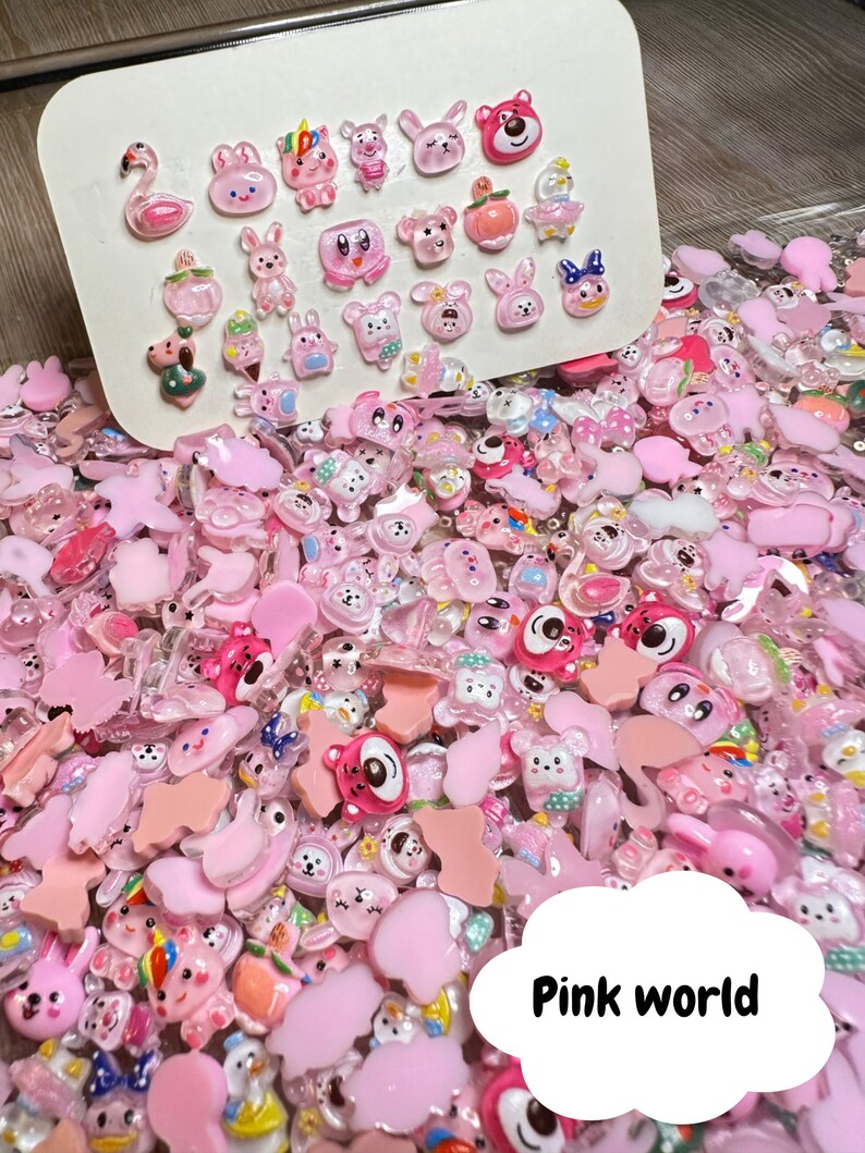 Pu&ograve; includere: Un vasto assortimento di ciondoli in plastica rosa e trasparente con vari animali e personaggi dei cartoni animati. I ciondoli sono sparsi su una superficie con un cartoncino bianco sullo sfondo che mostra una selezione pi&ugrave; piccola degli stessi ciondoli.