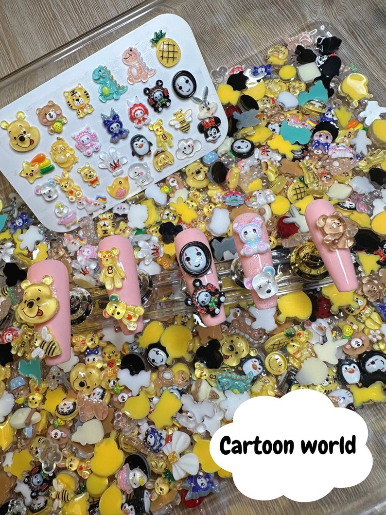 Pu&ograve; includere: Una variet&agrave; di ciondoli colorati con personaggi dei cartoni animati, tra cui Winnie the Pooh, un pinguino, un dinosauro, un'ape e un ananas. I ciondoli sono disposti su un vassoio bianco e una superficie rosa. Il testo "Cartoon World" &egrave; scritto in una forma di nuvola bianca.
