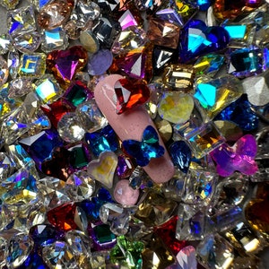 May include: Assortment of colourful rhinestones in various shapes, including hearts, squares, butterflies, and stars. The rhinestones are scattered on a grey surface.