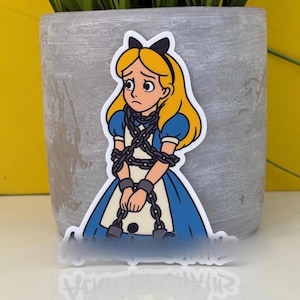 Op de afbeelding: Een sticker met een cartoonillustratie van Alice uit Alice in Wonderland, gebonden in kettingen. Alice wordt afgebeeld met blond haar, een blauwe jurk en een verdrietige uitdrukking. De sticker heeft een witte rand en staat voor een grijze pot.