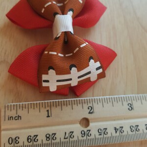 Pode incluir: Um la&ccedil;o de cabelo vermelho e marrom com um design de bola de futebol. O la&ccedil;o mede aproximadamente 2,5 cm de largura.