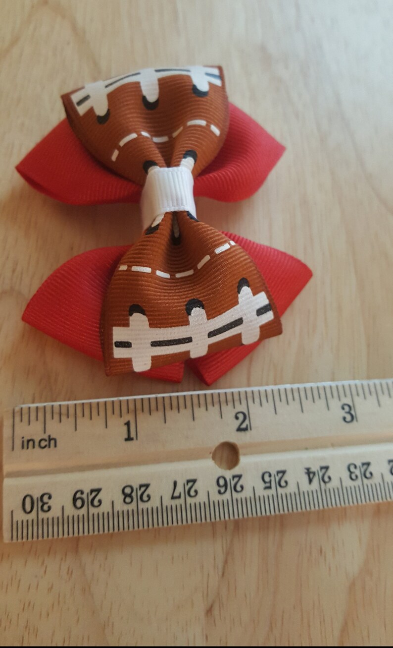 Pode incluir: Um la&ccedil;o de cabelo vermelho e marrom com um design de bola de futebol. O la&ccedil;o mede aproximadamente 2,5 cm de largura.