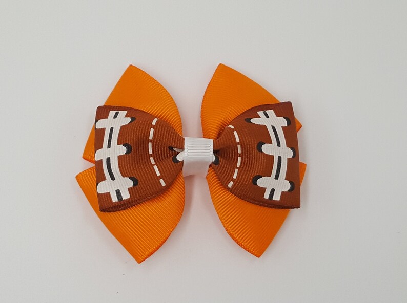Pode incluir: La&ccedil;o de cabelo laranja e marrom com um design de bola de futebol. O la&ccedil;o &eacute; amarrado com uma fita branca.