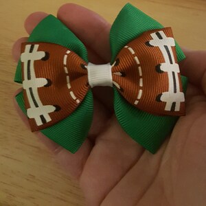Pode incluir: Um la&ccedil;o de cabelo verde e marrom com um design de bola de futebol. O la&ccedil;o &eacute; feito de fita e tem um centro branco.