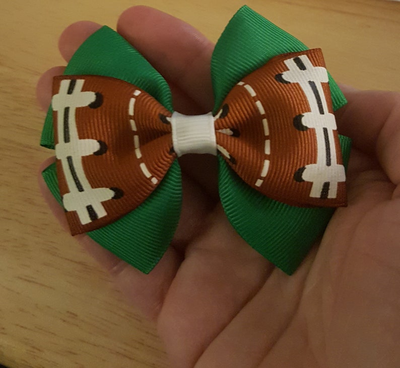 Pode incluir: Um la&ccedil;o de cabelo verde e marrom com um design de bola de futebol. O la&ccedil;o &eacute; feito de fita e tem um centro branco.
