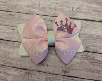 May include: A light pink hair bow with a glittery white base. The bow features the word "Toss" in white script on the pink ribbon. A glittery crown design is on the top right side of the bow.
