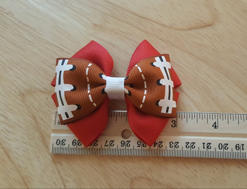 Pode incluir: Um la&ccedil;o de cabelo vermelho e marrom com um design de bola de futebol. O la&ccedil;o tem aproximadamente 2,5 polegadas de largura e &eacute; medido contra uma r&eacute;gua.