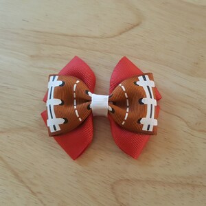 Pode incluir: Um la&ccedil;o de cabelo vermelho e marrom com um design de bola de futebol. O la&ccedil;o &eacute; feito de fita e tem um centro branco.
