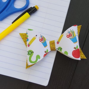 May include: A white bow with a gold glitter trim and a school-themed print featuring pencils, apples, and a worm with glasses.