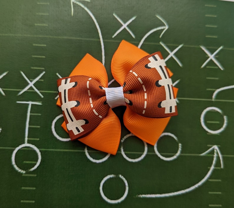 Pode incluir: Um la&ccedil;o de cabelo marrom e laranja com um design de bola de futebol. O la&ccedil;o tem um centro branco e &eacute; amarrado com uma fita branca.