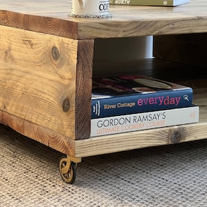May include: A rustic wooden coffee table with a lower shelf. The table has a natural wood finish and is on wheels. Two cookbooks are on the lower shelf: "River Cottage everyday" and "Gordon Ramsay's Ultimate Cookery Course."