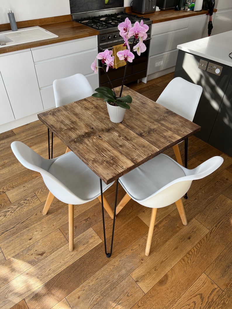 May include: A square wooden dining table with black hairpin legs and four white chairs. A potted pink orchid sits in the center of the table.