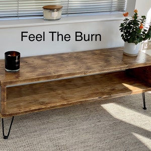 Puede incluir: Un soporte de TV de madera r&uacute;stica con un estante y patas de horquilla. El soporte tiene un acabado marr&oacute;n desgastado y el texto "Feel The Burn" est&aacute; escrito en la pared detr&aacute;s de &eacute;l.