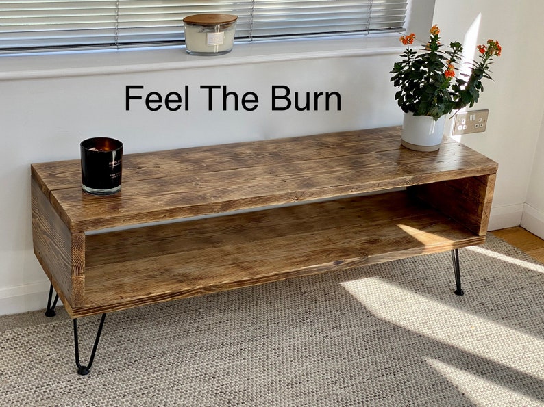 Puede incluir: Un soporte de TV de madera r&uacute;stica con un estante y patas de horquilla. El soporte tiene un acabado marr&oacute;n desgastado y el texto "Feel The Burn" est&aacute; escrito en la pared detr&aacute;s de &eacute;l.
