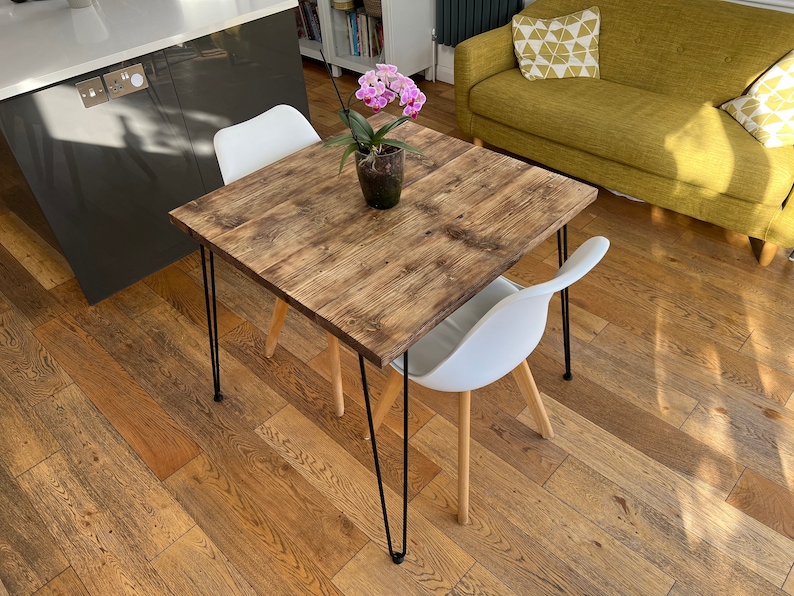 May include: A square wooden table with a rustic finish and black hairpin legs. The table is set with a white chair and a potted orchid plant.