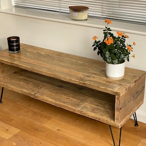 Puede incluir: Un soporte de TV de madera r&uacute;stica con un estante, con un acabado desgastado y patas de horquilla negras. El soporte est&aacute; decorado con una planta en maceta con flores naranjas y una vela.