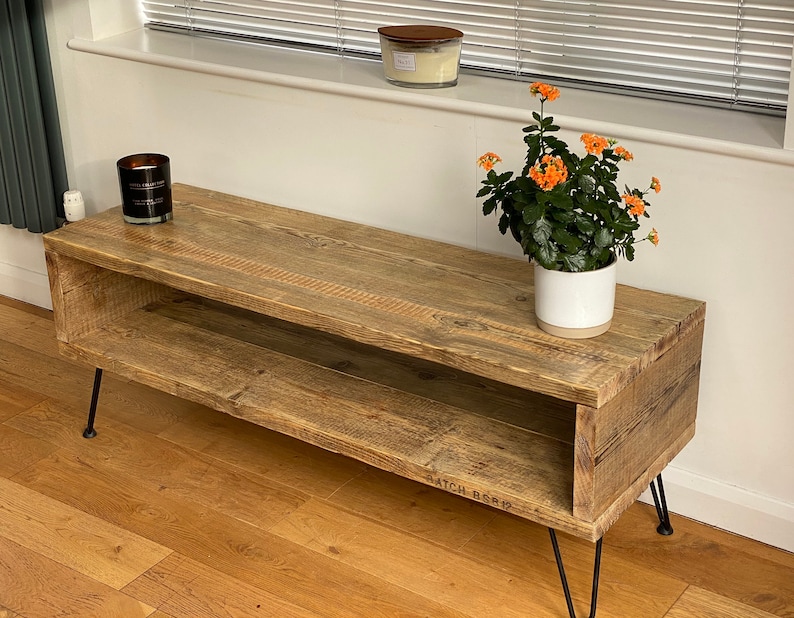 Puede incluir: Un soporte de TV de madera r&uacute;stica con un estante, con un acabado desgastado y patas de horquilla negras. El soporte est&aacute; decorado con una planta en maceta con flores naranjas y una vela.