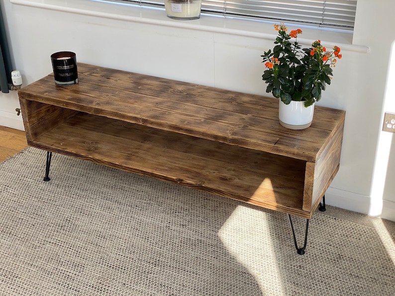 Puede incluir: Un soporte de TV de madera r&uacute;stica con un estante y patas de horquilla negras. El soporte est&aacute; sobre una alfombra gris claro y tiene una planta en maceta con flores naranjas en la parte superior.
