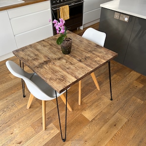 May include: A square wooden table with black hairpin legs and a light wood finish. The table is set with a white chair and a potted orchid with pink flowers.