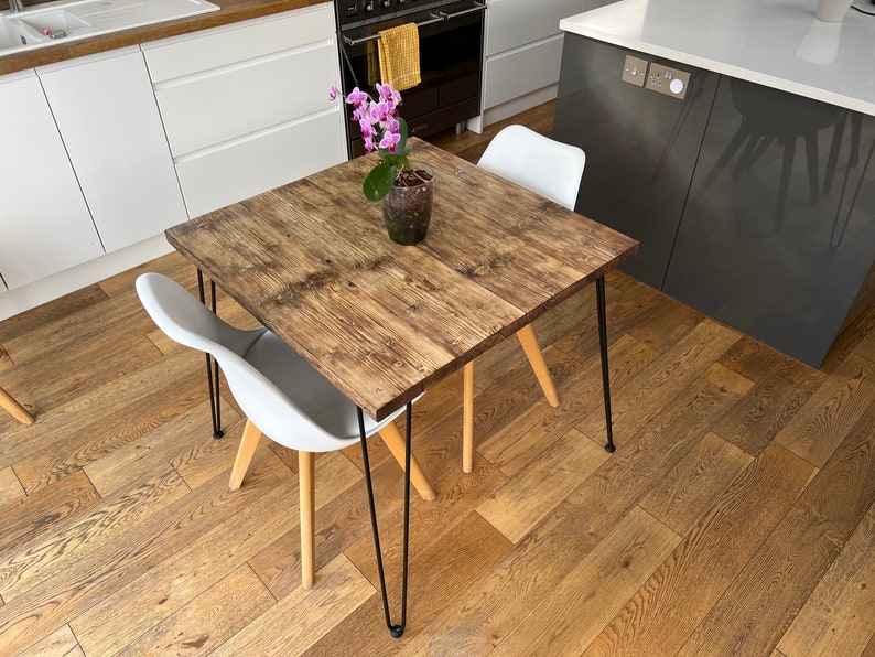 May include: A square wooden table with black hairpin legs and a light wood finish. The table is set with a white chair and a potted orchid with pink flowers.