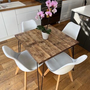 May include: A square wooden dining table with black hairpin legs and four white chairs. A potted orchid with pink flowers sits in the center of the table.